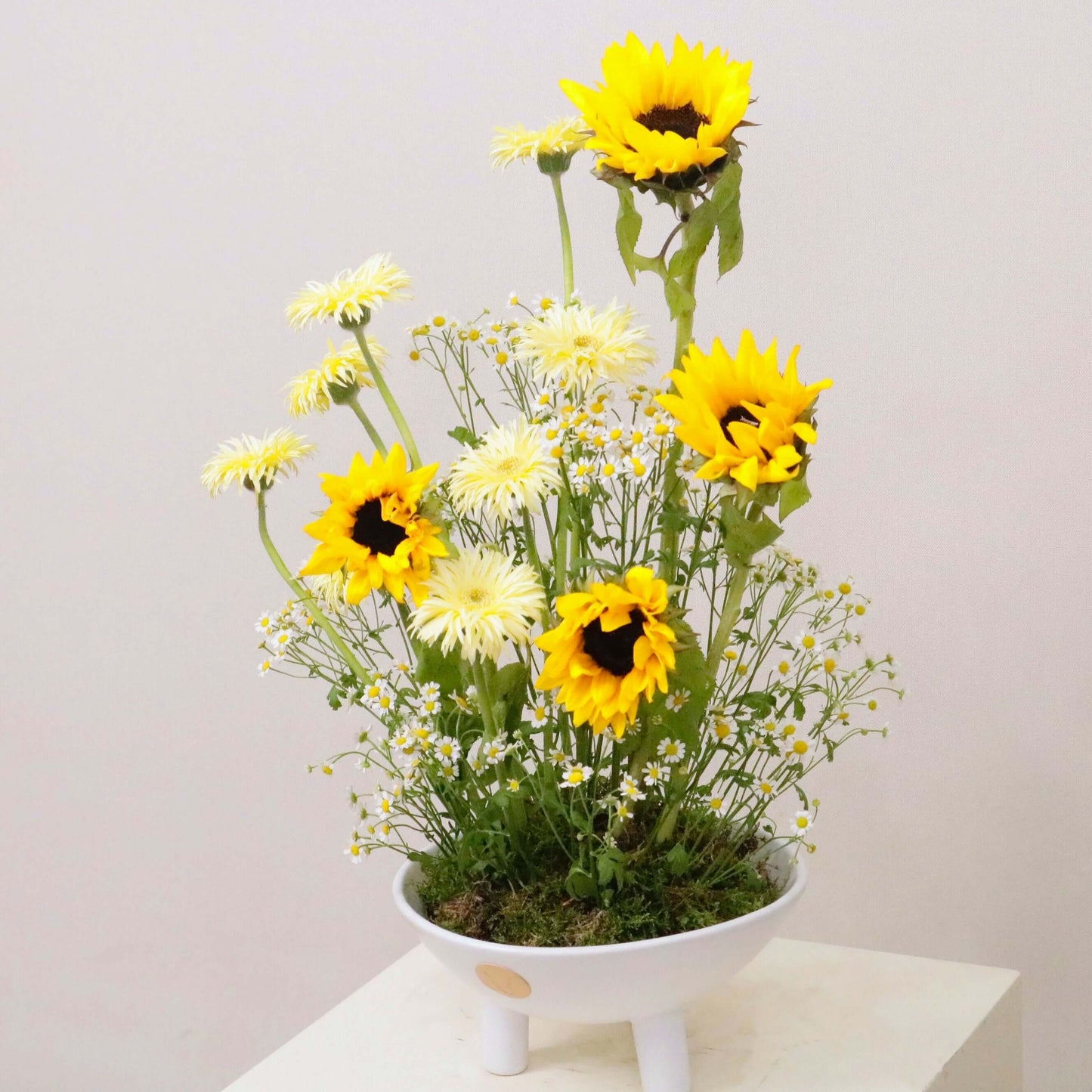 Aestas floral arrangement with sunflowers, yellow gerbera daisies, and matricaria in a white vase, symbolizing summer's warmth.