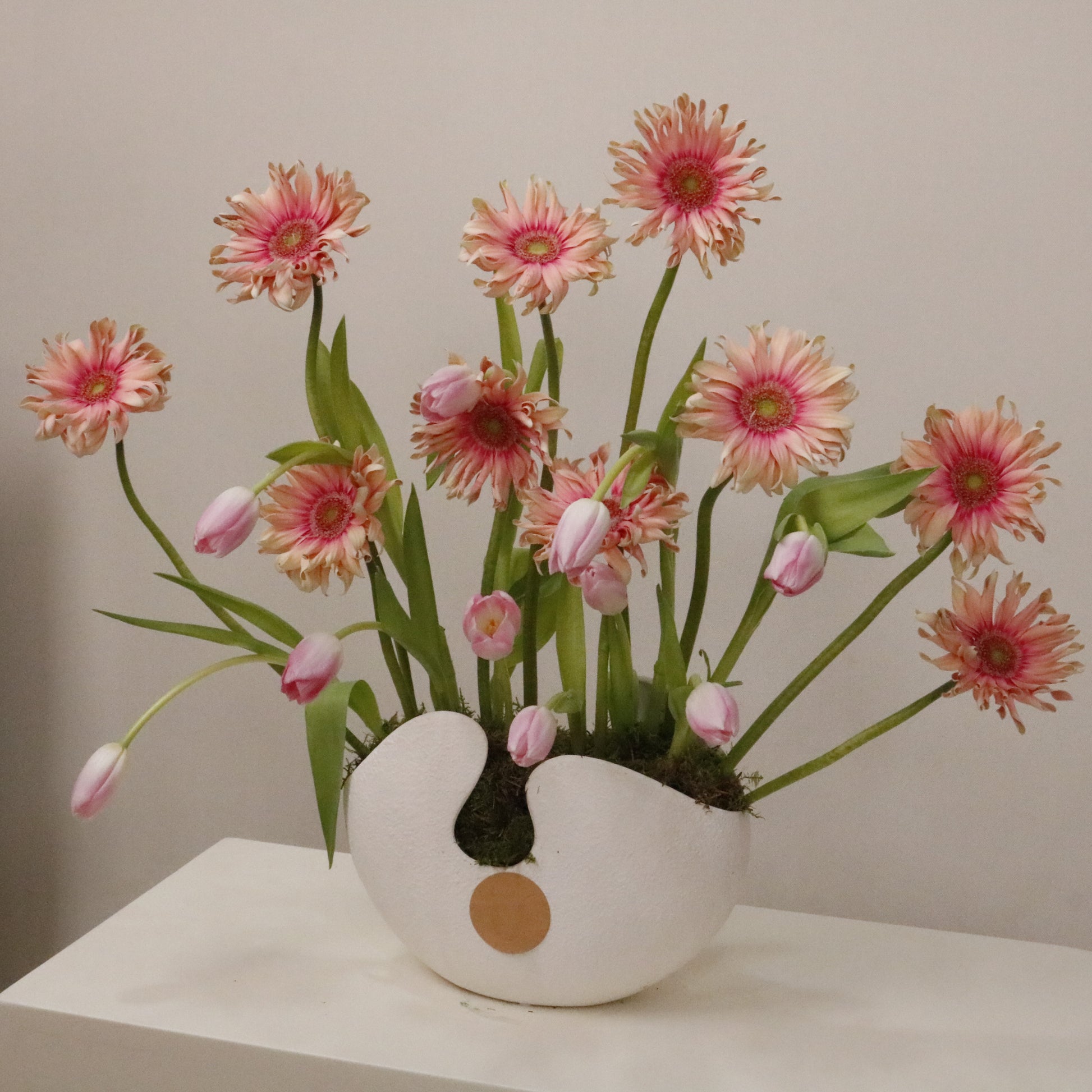 The perfect artistic luxury gift featuring pink gerberas and pastel tulips in a white ceramic vase.