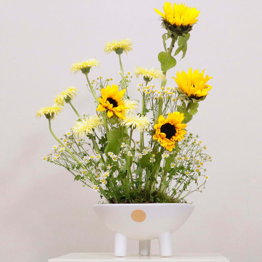 Aestas floral arrangement with sunflowers, yellow gerbera daisies, and matricaria in a white vase, bringing summer warmth and brightness.
