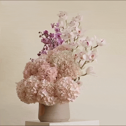 A floral arrangement of pink hydrangeas, pink orchids, and purple cymbidium, inspired by a famous Italian song, in a beige vase.